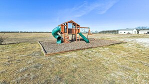 A playground with a wooden house structure, slides, and a swing set in an open field. - 8 Mi to White Rock Lake: Spacious Krum Home (Krum)