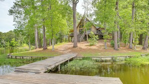 A cabin surrounded by tall trees and a wooden dock extending into a calm lake. - Riverfront Shelby Home w/ Private Boat Dock! (Shelby)