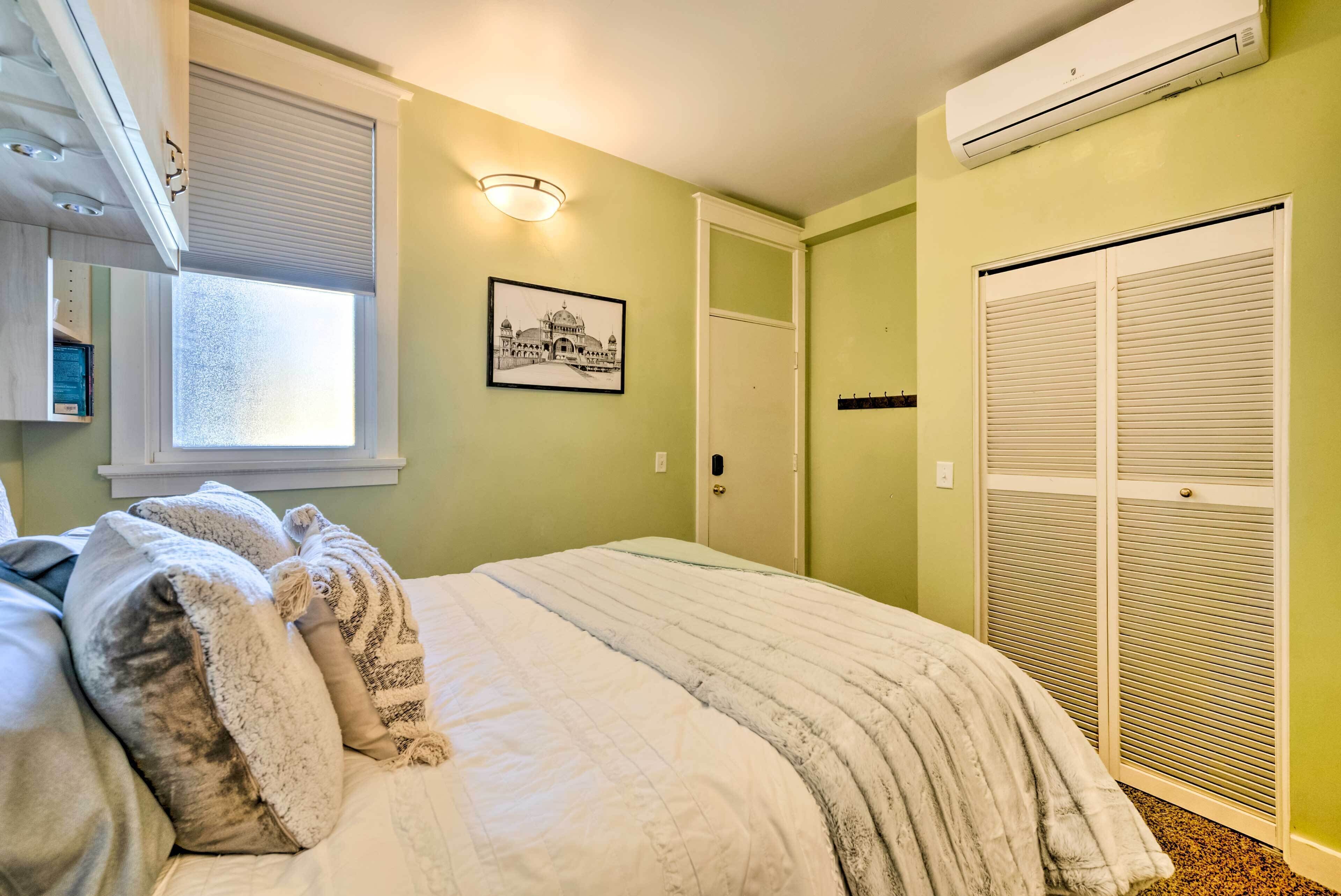 A bedroom with a bed, a window with blinds, a wall-mounted air conditioner, a door with a closet, and a framed picture on the wall.
