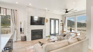 A modern living room with a stone fireplace, a flat-screen TV, a large sofa, and a dining area with a bar. - Romantic Mountain Retreat w/ Scenic Views! (Idaho Springs)