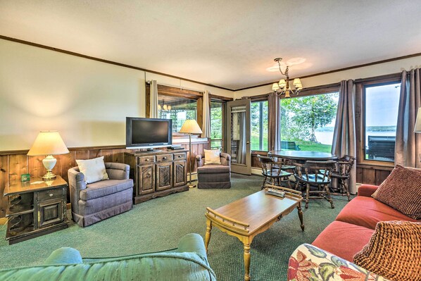 A spacious living room with a television, wooden furniture, a dining area, and large windows offering a view of the outdoors.