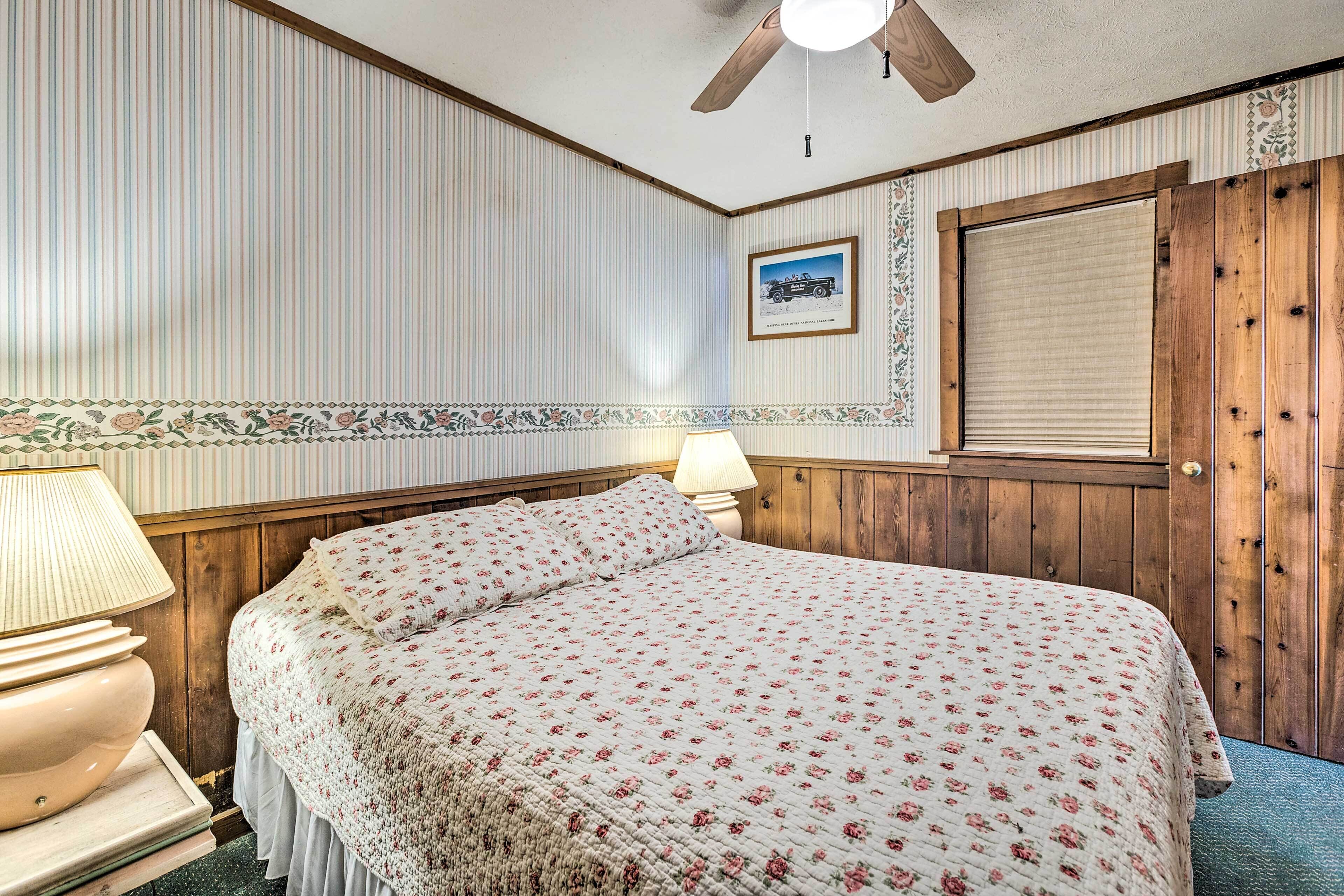 Ein Schlafzimmer mit einem Bett, einem Holz-Kopfteil, einem Deckenventilator, einem Fenster mit Jalousien und einem gerahmten Bild an der Wand.