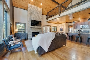 A spacious living room with a fireplace, a large sofa, and a kitchen area with bar stools. - Serene Broken Bow Cabin w/ Hot Tub & Fire Pit! (Broken Bow)