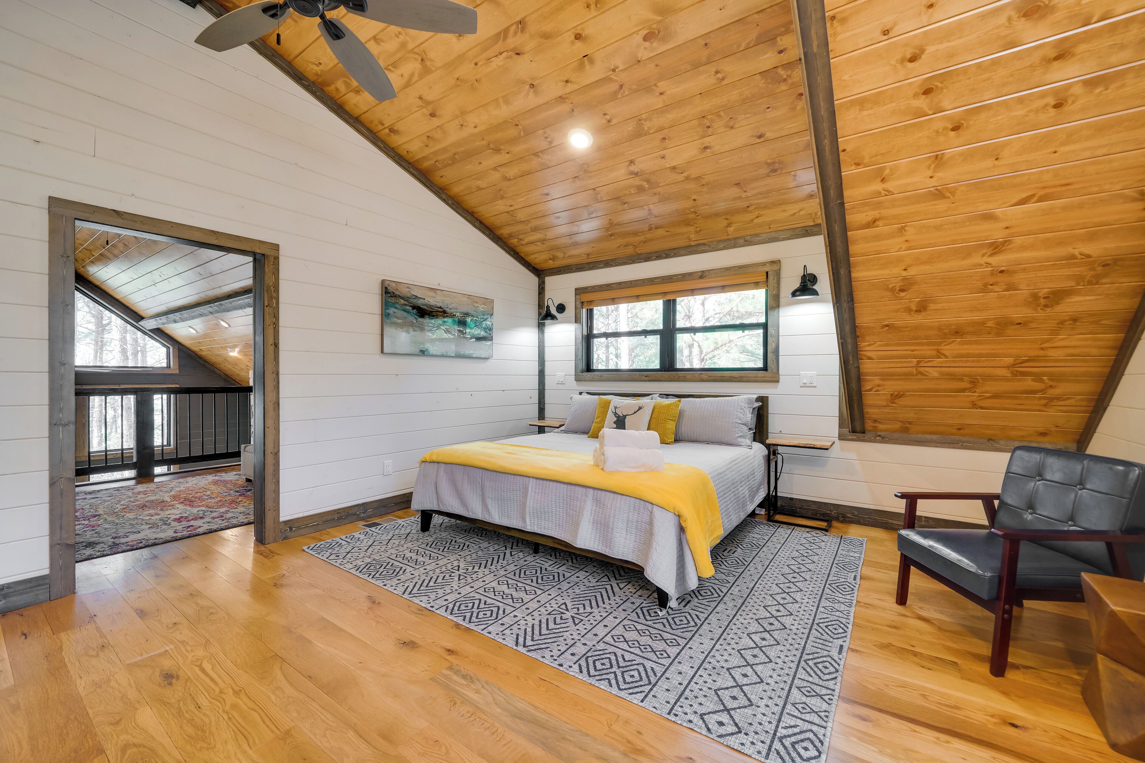 A bedroom with a wooden ceiling, a bed with a yellow blanket, a black and white patterned rug, and a window with a view of trees.