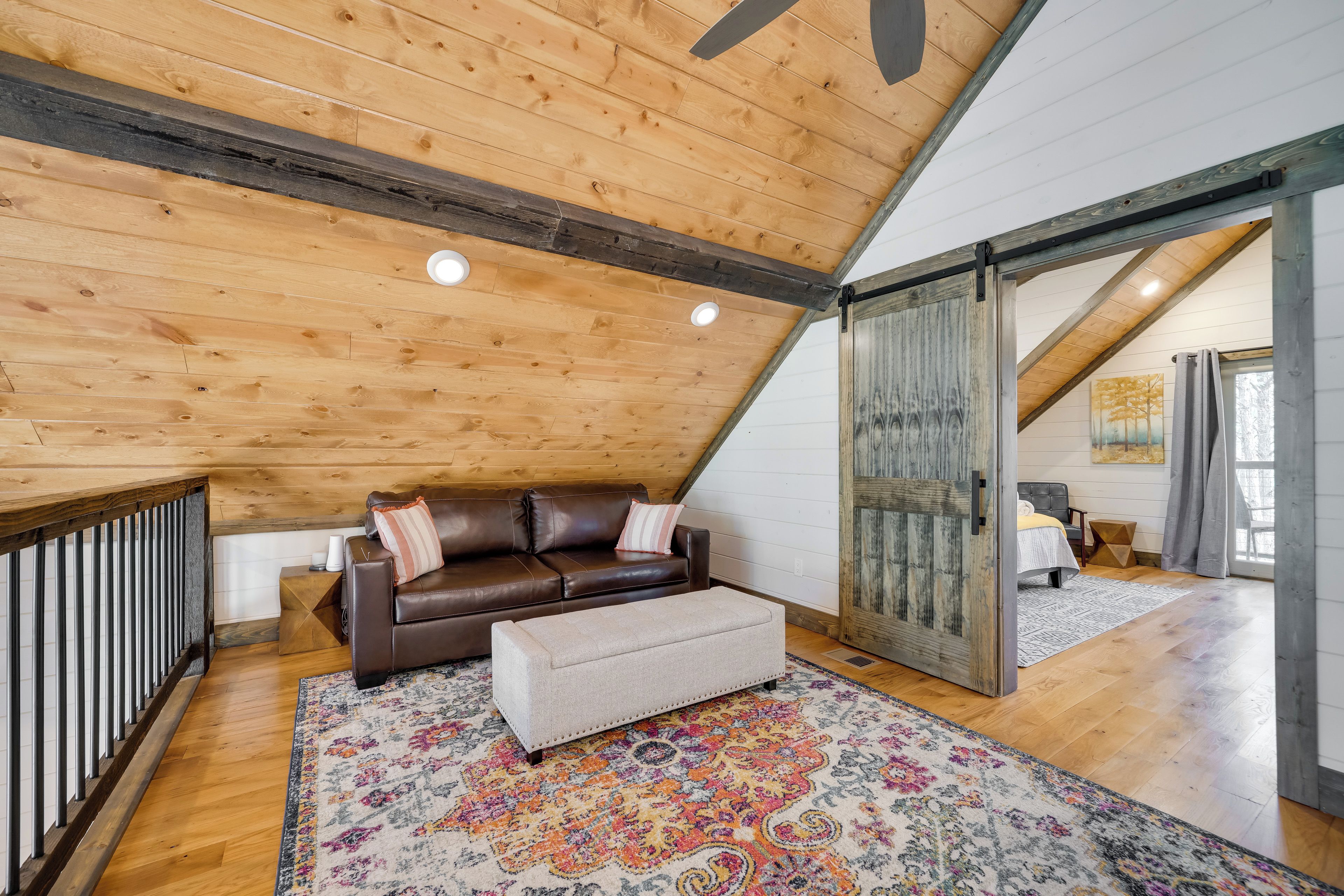 A cozy loft bedroom with a wooden ceiling, a brown leather sofa, a matching ottoman, and a colorful rug.