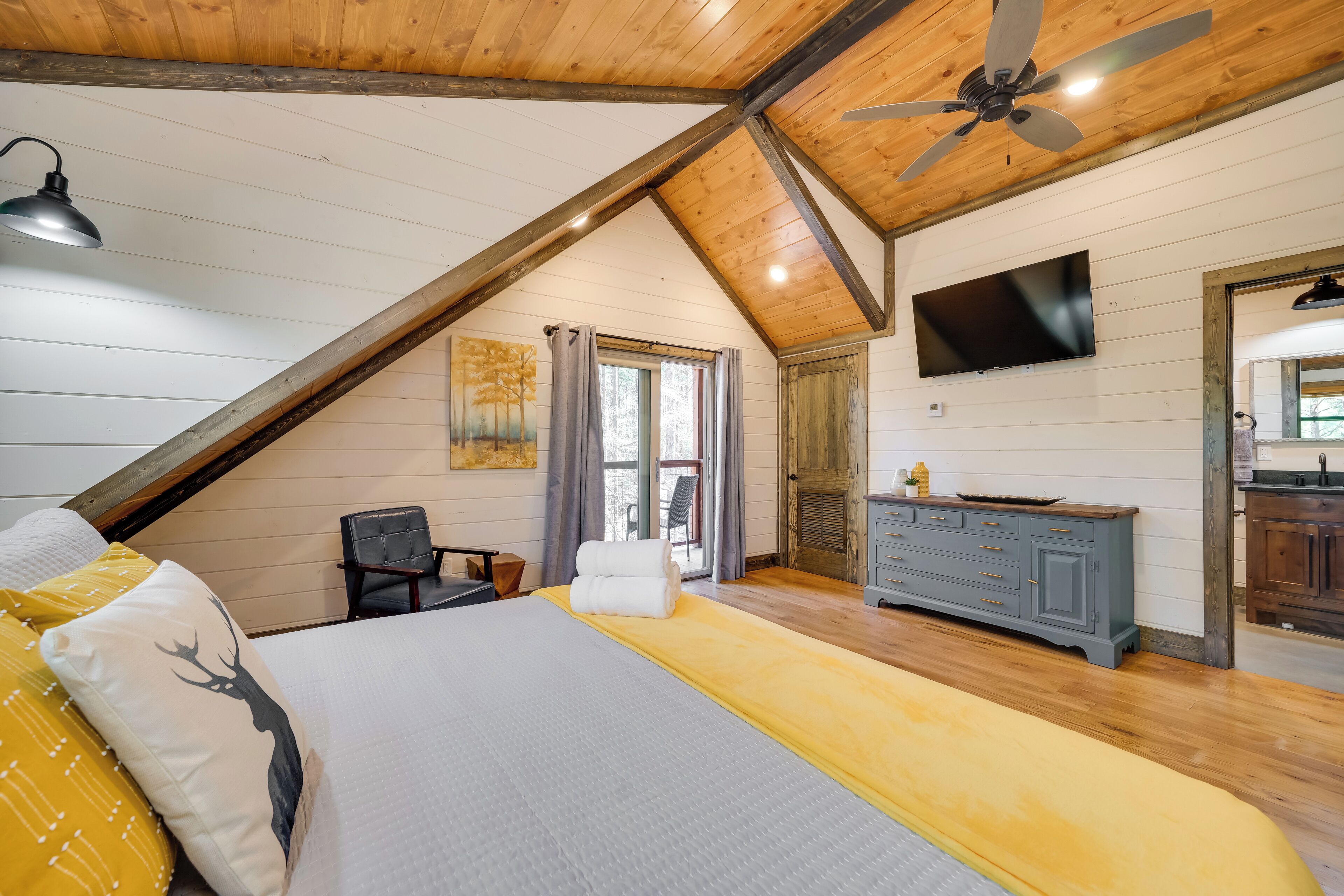A bedroom with a sloped ceiling, a bed with a white and yellow bedspread, a gray headboard, a desk, a chair, a television, and a wooden dresser.
