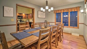 Zona pranzo con tavolo e sedie in legno, un lampadario e una cucina visibile attraverso un'apertura.