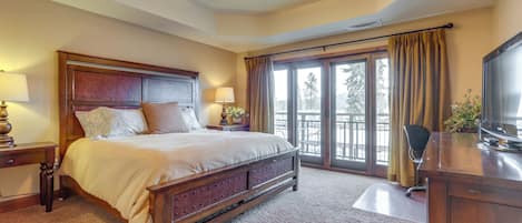 A bedroom with a large bed, two bedside tables with lamps, a desk with a television, and a view of the outdoors through a sliding glass door.