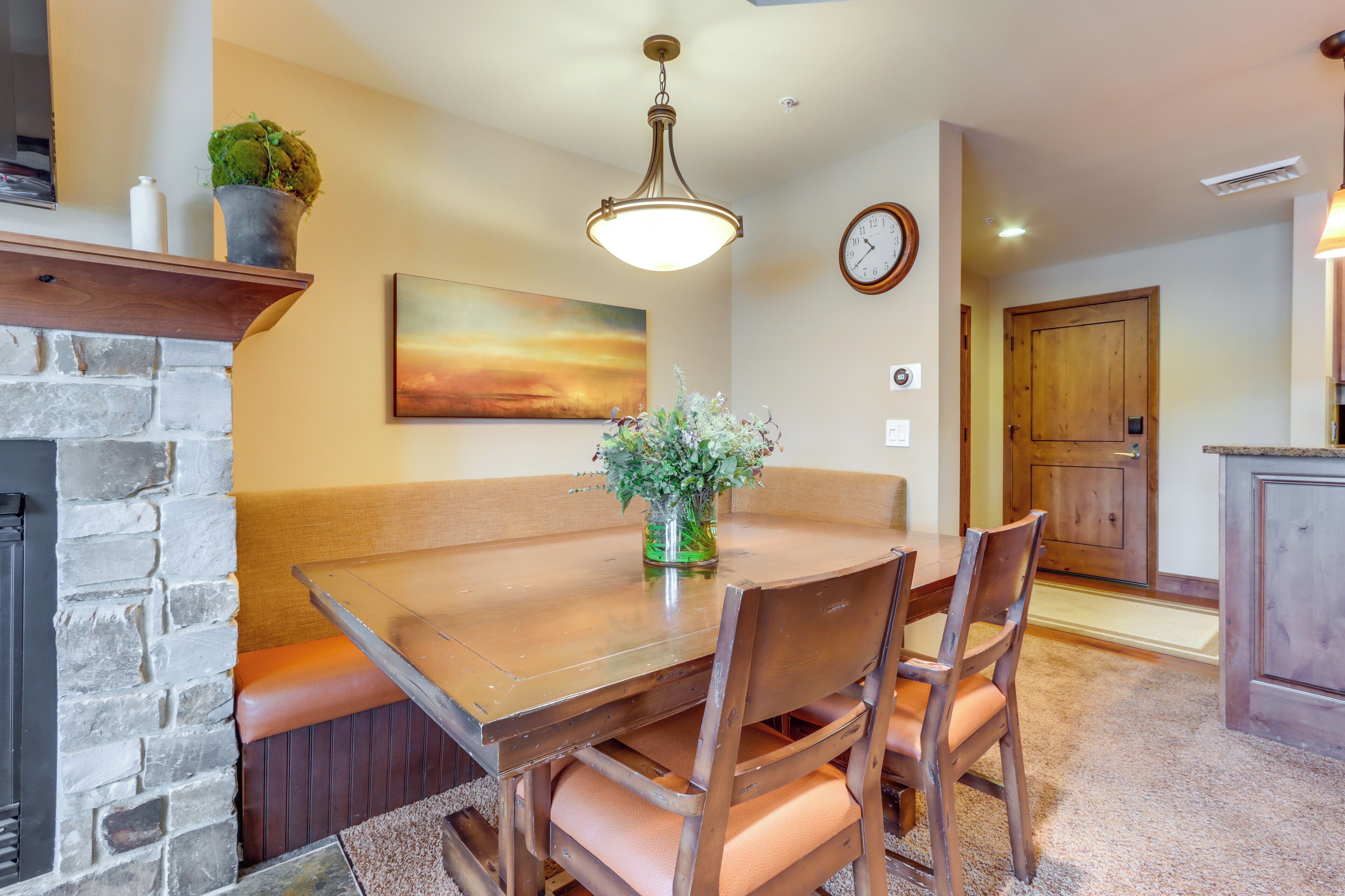 A dining area with a wooden table, cushioned bench seating, a vase with flowers, a fireplace, a painting on the wall, and a clock.