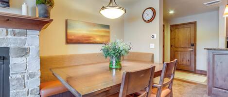A dining area with a wooden table, cushioned bench seating, a vase with flowers, a fireplace, a painting on the wall, and a clock.