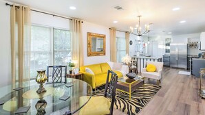A modern living room with a yellow sofa, a glass dining table, and a kitchen in the background. - Lovely Historic Fort Worth Home w/ Deck (Fort Worth)