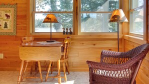 Interior de una cabaña de madera con una mesa de comedor, sillas, un sillón de mimbre, dos lámparas y una ventana con vista a árboles.
