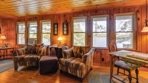 Interior acogedor de una cabaña con paredes de madera, ventanas amplias, dos sillones, una mesa de comedor y una lámpara.