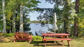 Un área de picnic junto al lago, con un banco de madera y una mesa de picnic, rodeada de árboles altos y una vista de un bote en el agua.