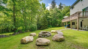 Une maison avec un foyer en pierre entouré de gros rochers, un patio couvert avec des sièges d’extérieur et un paysage boisé en arrière-plan.