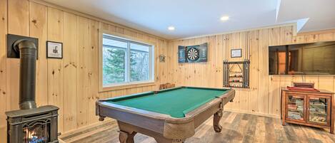 A pool table in a room with wooden walls, a large window, and a fireplace.