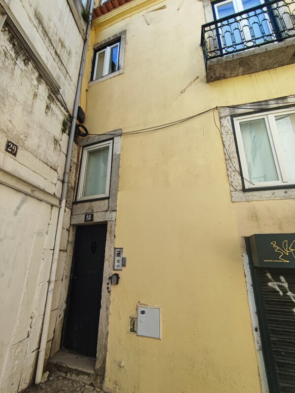 A narrow alleyway with a yellow building, a black door, and a small window with a white frame.