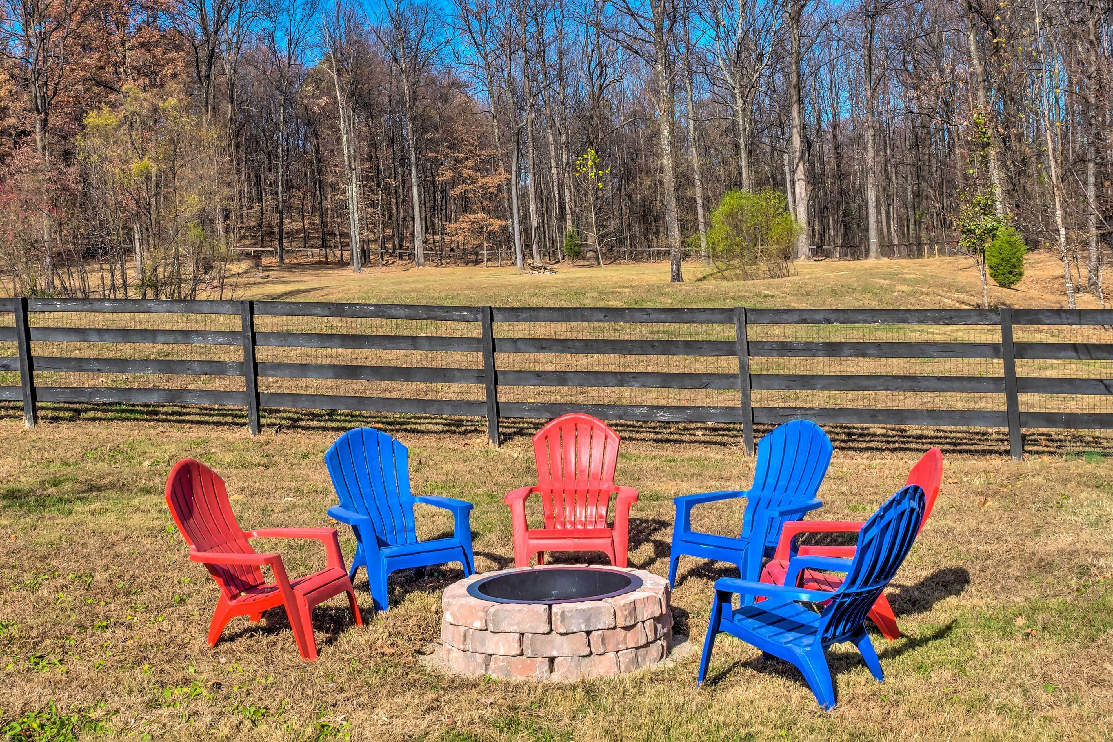 A fire pit surrounded by four Adirondack chairs in red and blue, set against a backdrop of a fenced field and trees.
