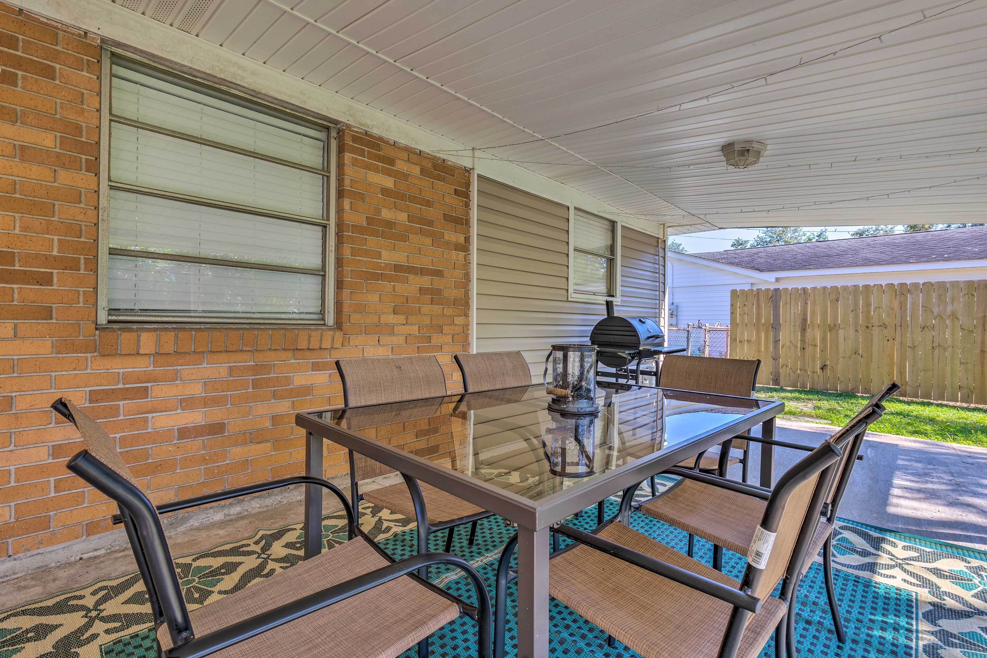 A covered patio with a glass-top table and six chairs, a barbecue grill, and a wooden fence in the background.