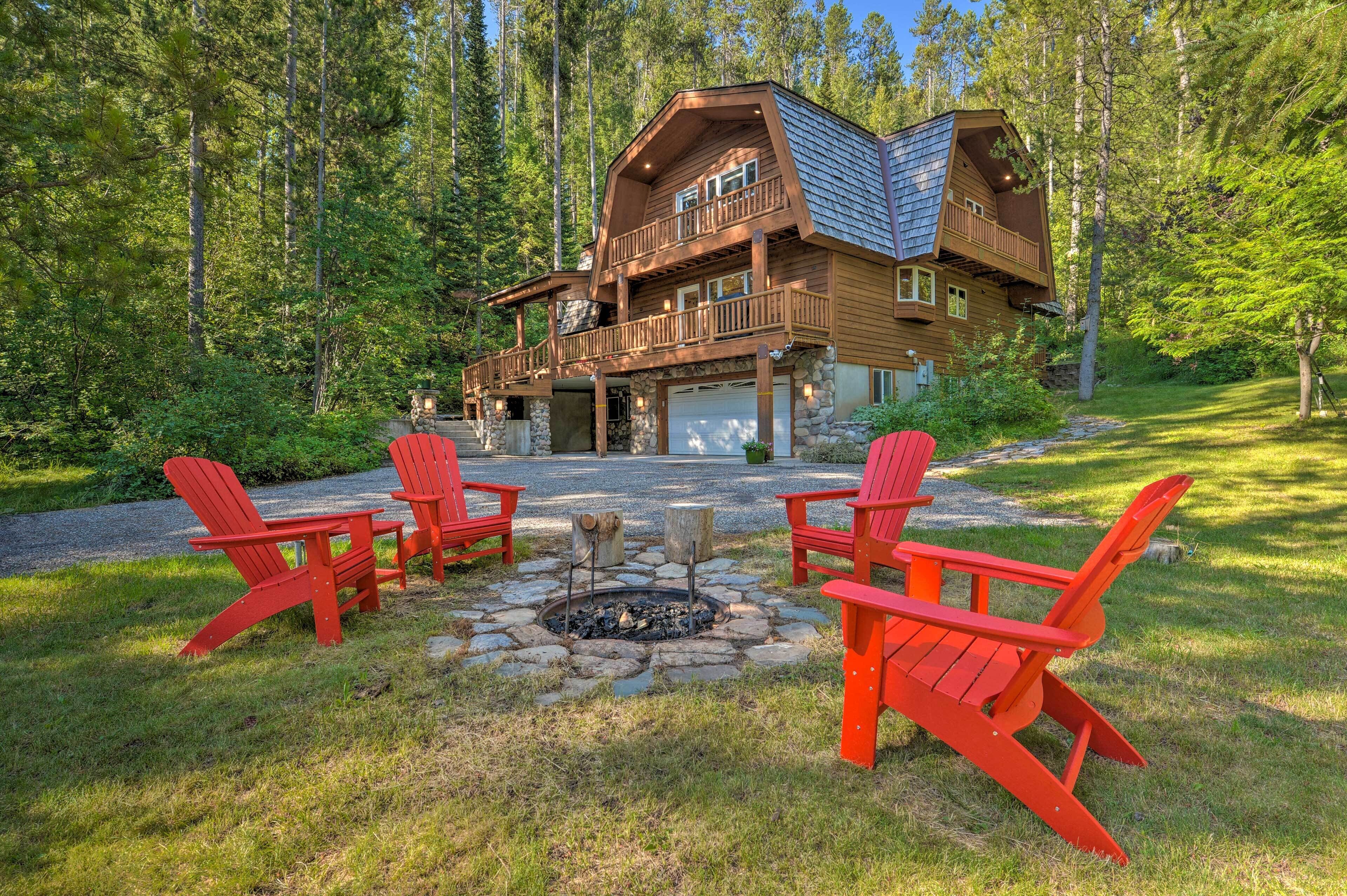 A cabin with a stone fire pit and red Adirondack chairs in a forest setting.