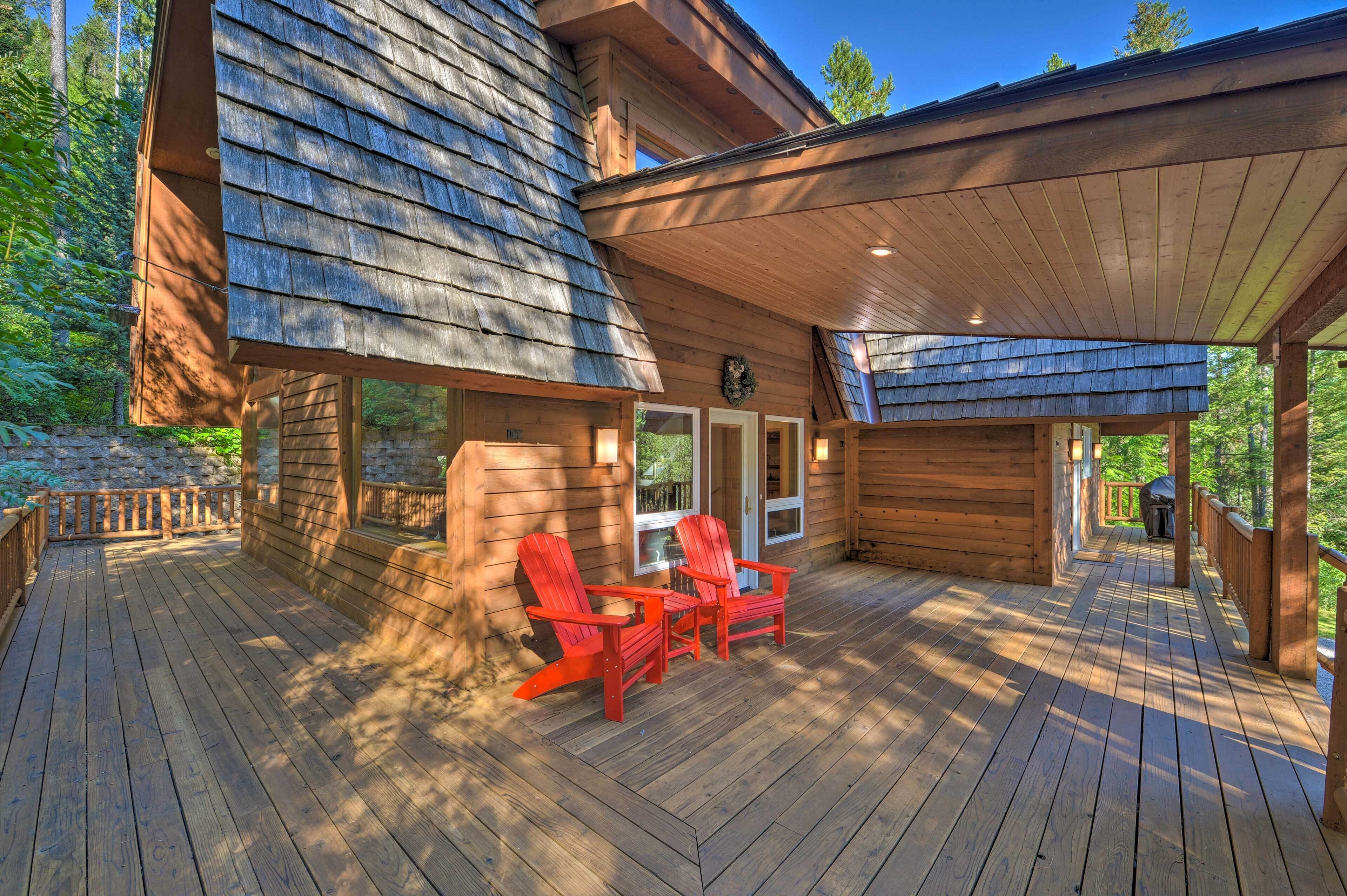 A wooden deck with red Adirondack chairs and a table, surrounded by a stone wall and trees.