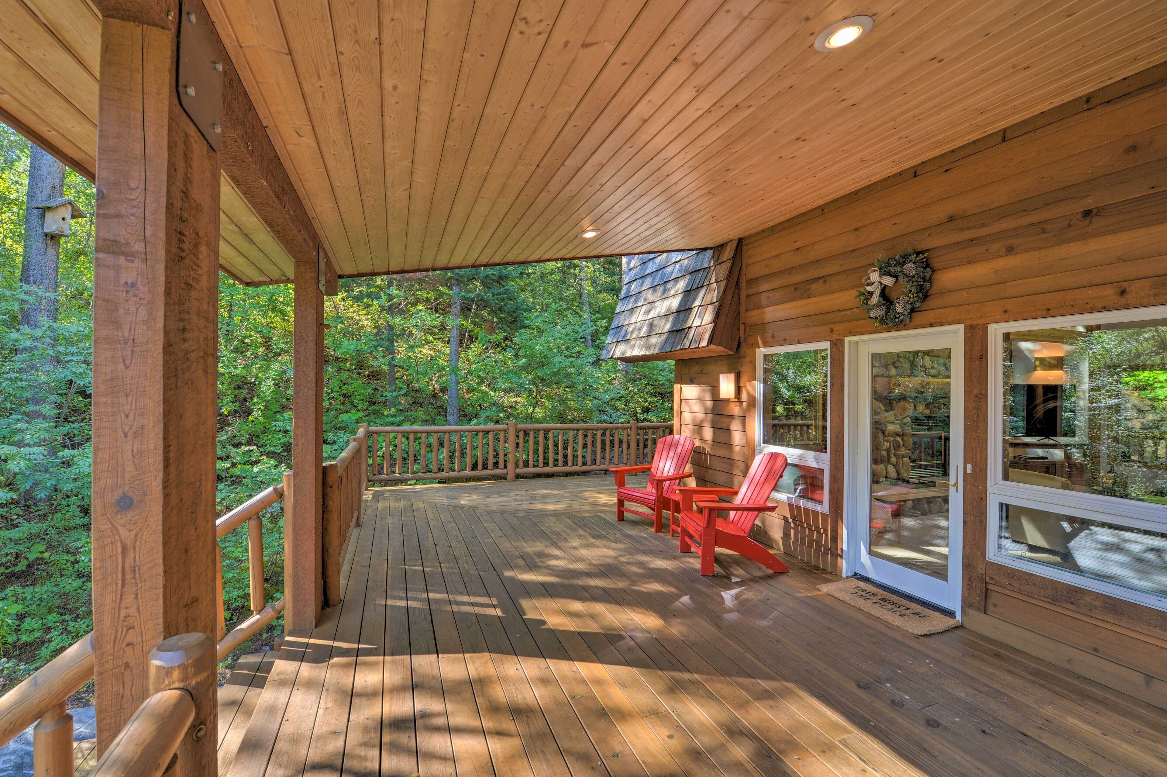 A wooden deck with red Adirondack chairs, a stone fireplace, and a view of the forest.