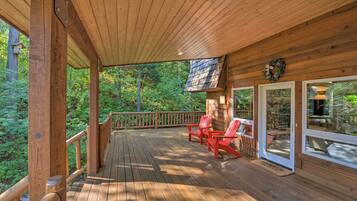 A wooden deck with red Adirondack chairs, a stone fireplace, and a view of the forest.