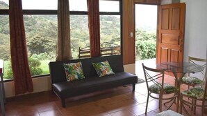 A room with a sofa, a dining table with chairs, and large windows offering a view of greenery. - Apartments Monte Luna Mountain View (Monteverde)
