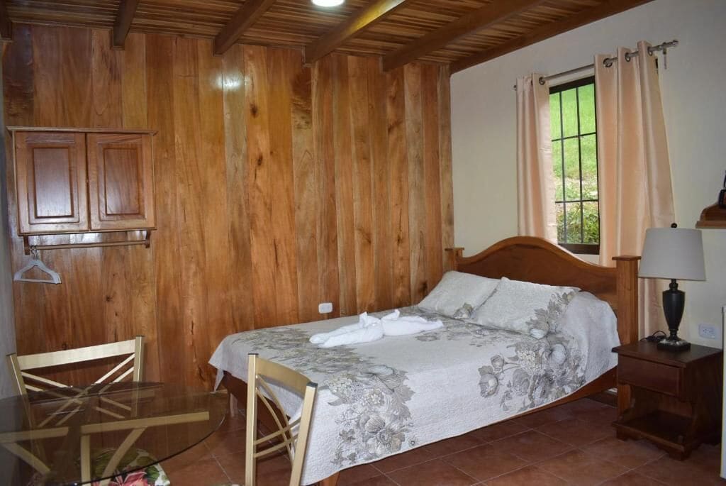 A wooden-paneled bedroom with a bed, a bedside table, a lamp, a chair, and a small table.