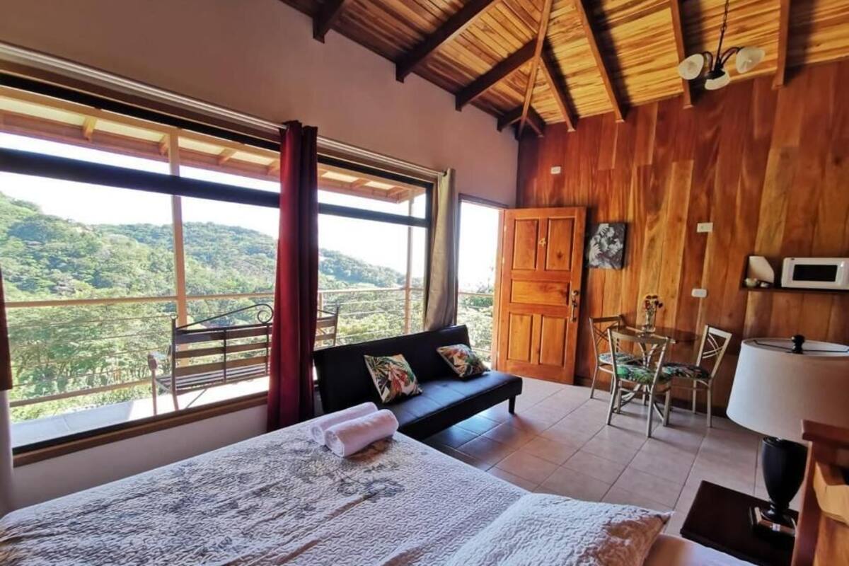 A bedroom with a large bed, a sofa, a dining table, and a view of the outdoors.