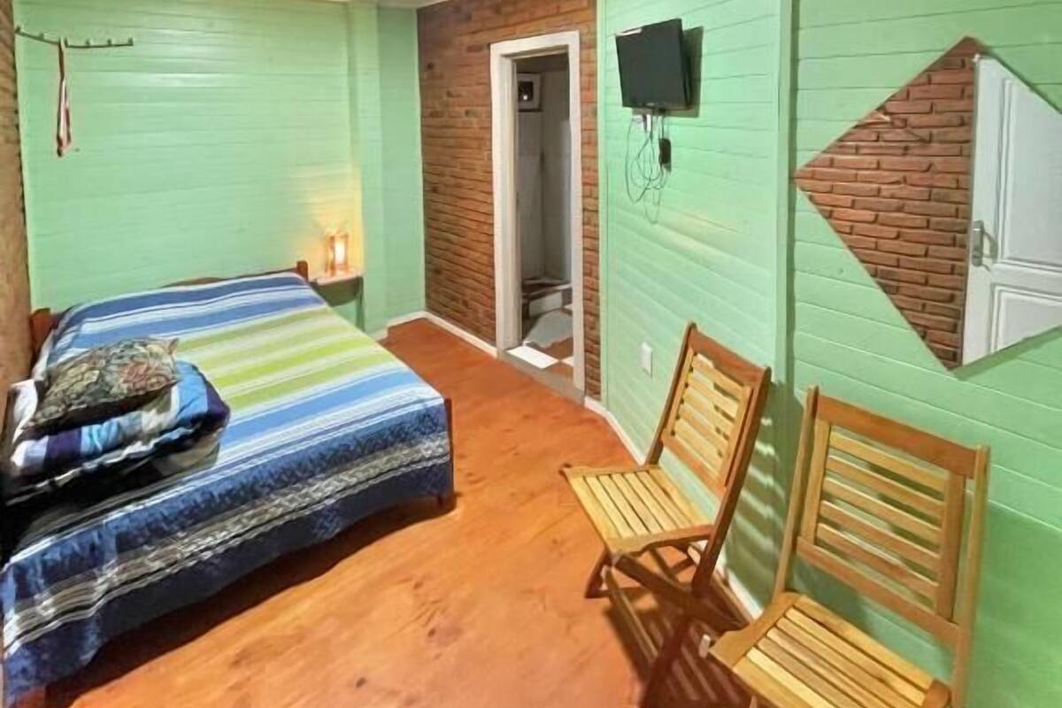 Una habitación con una cama, dos sillas de madera, un televisor montado en la pared y un pequeño muro decorativo de ladrillo.