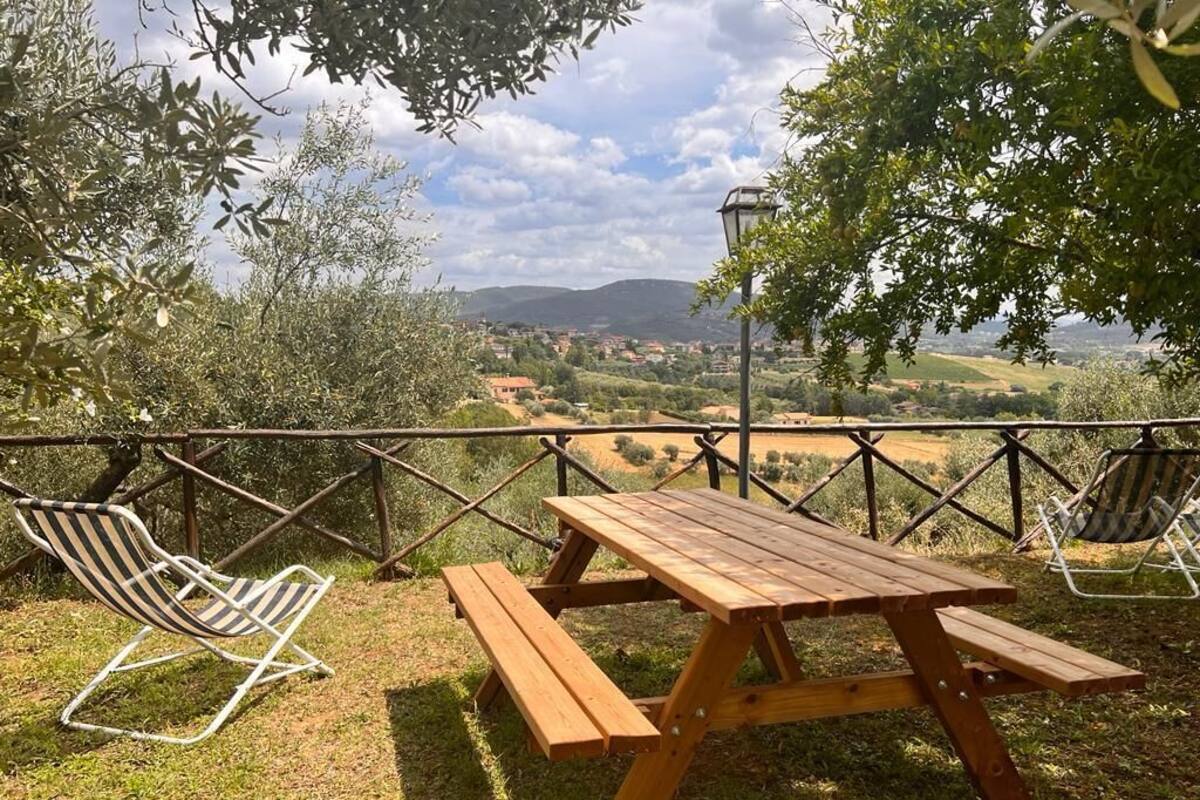 Mesa de piquenique com duas cadeiras, cercada por uma vedação metálica, com vista para uma paisagem pitoresca com colinas e uma cidade ao longe.