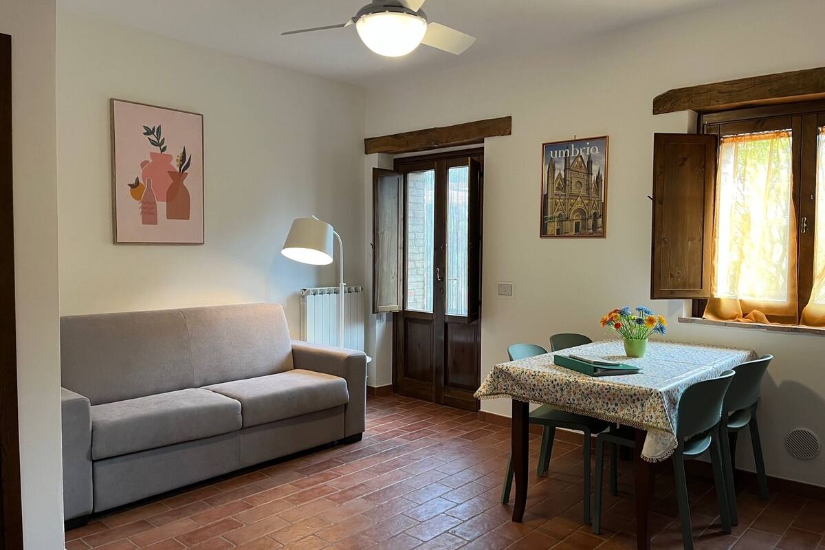 Uma sala de jantar com mesa posta para quatro pessoas, um sofá, um ventilador de teto e um radiador.