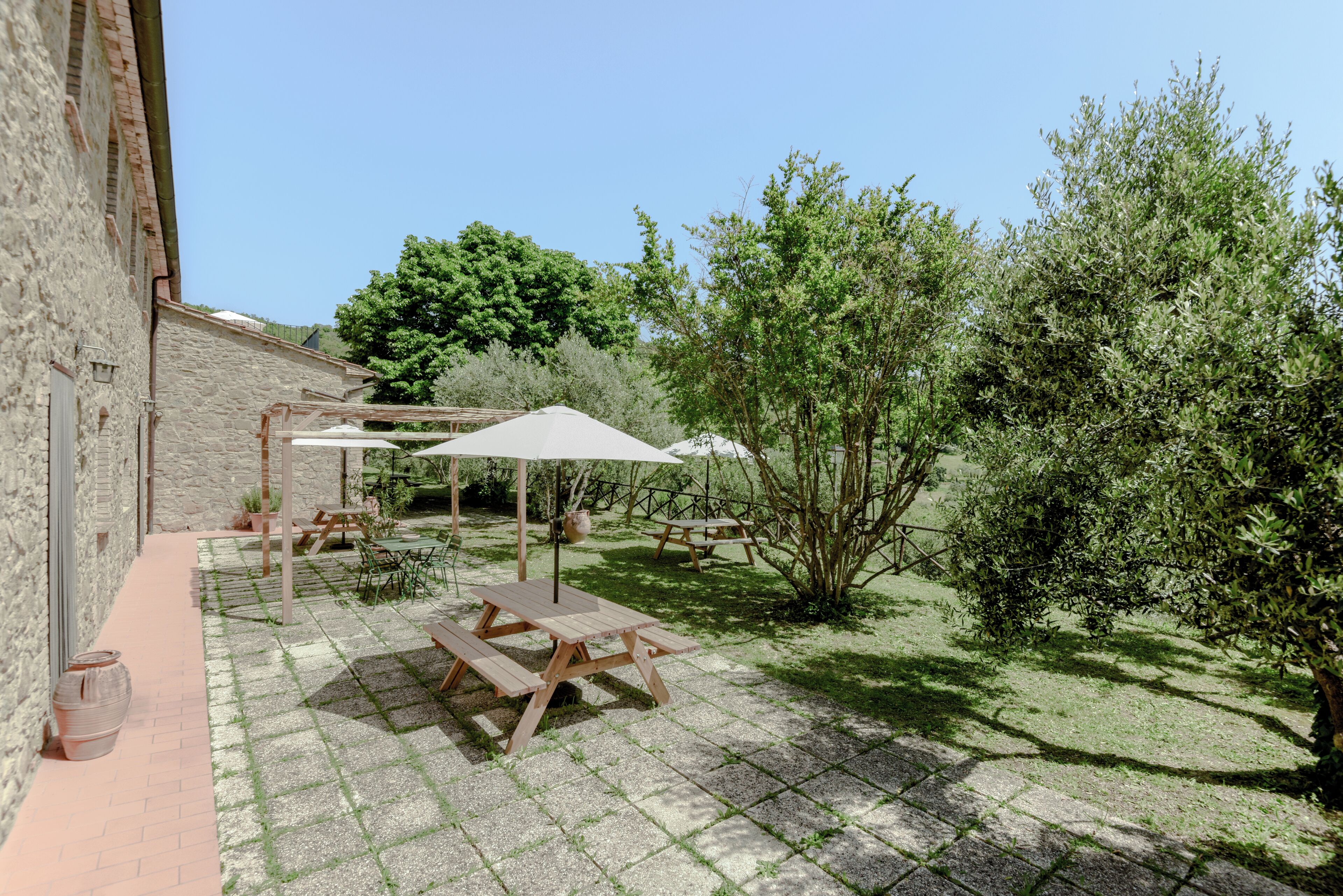 A patio area with picnic tables and umbrellas, surrounded by greenery and a stone building.