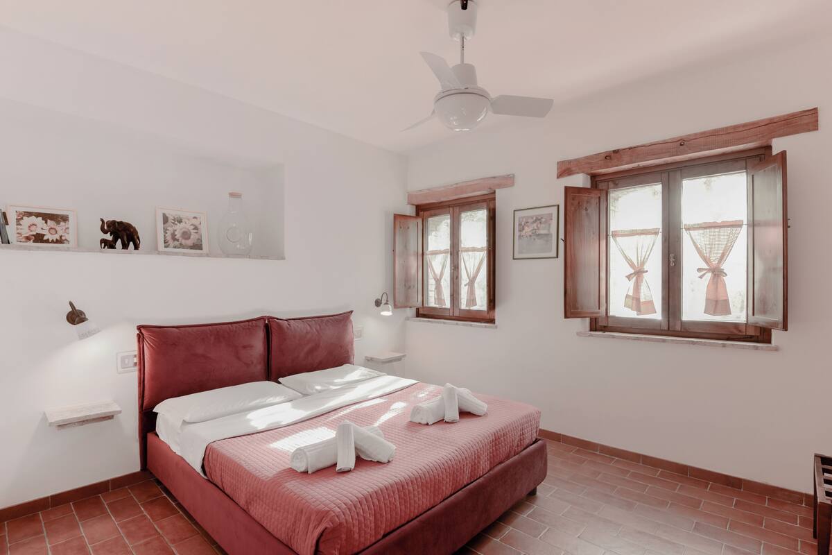 Quarto com cama, ventilador de teto, janela com cortinas e obra de arte na parede.