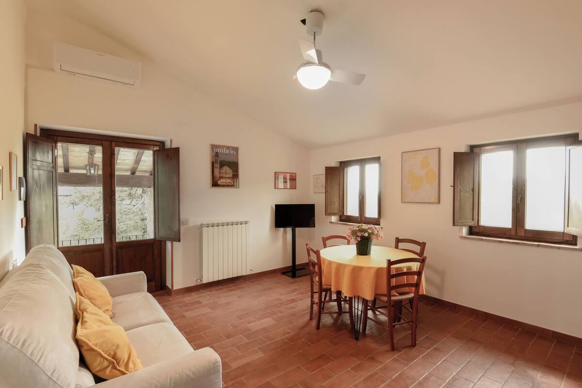 Uma sala de estar com sofá, uma mesa de jantar com cadeiras, um ventilador de teto e uma janela com vista.