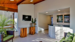 A modern interior with a wooden ceiling, a glass partition, a flat-screen TV, a reception desk, and a green sofa. - Hotel Encantos De Itaperapuã (Porto Seguro)