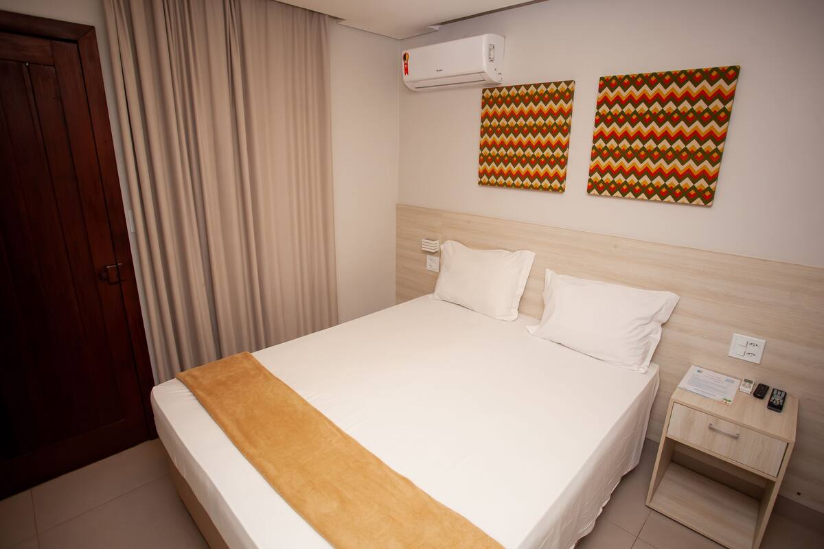 Una camera d'albergo con un letto grande, un comodino in legno, un condizionatore d'aria e due decorazioni murali a motivi decorati.