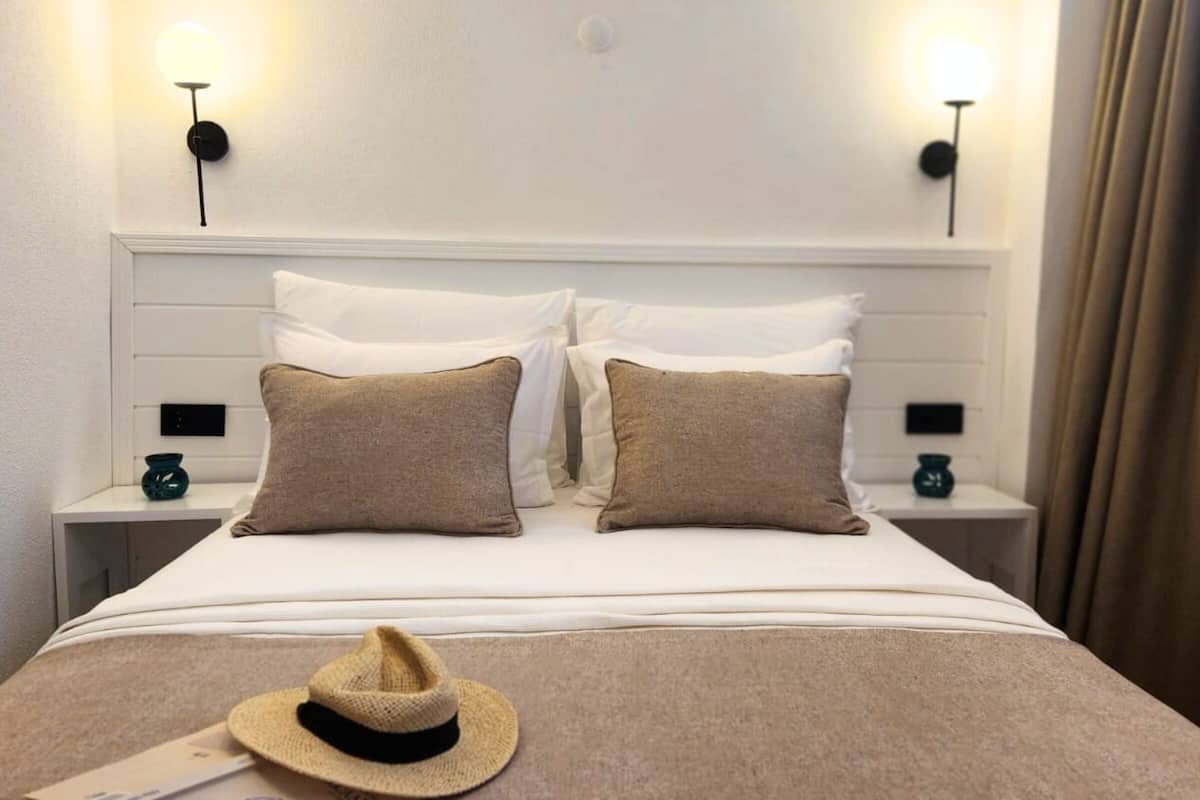 Una cama bien hecha con ropa blanca y dos almohadas marrones, un sombrero de paja sobre la cama, y dos mesitas de noche con lámparas.
