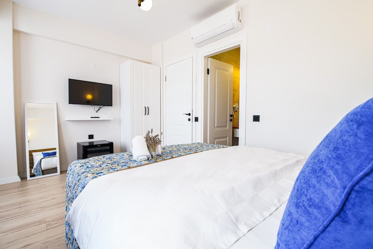 Una habitación de hotel con una cama, un televisor montado en la pared, una mesita de noche con lámpara y una puerta que da a otra habitación.