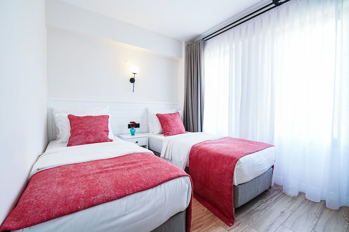 Habitación de hotel con dos camas, ropa de cama blanca, detalles en rojo, una mesita de noche con un florero y una ventana con cortinas traslúcidas.