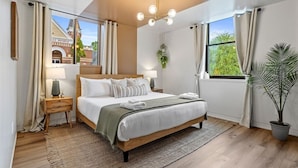 A modern bedroom with a large bed, bedside tables, a hanging light fixture, and a view of a building outside. - Alchemist @ The Vestry on Read (Columbia)