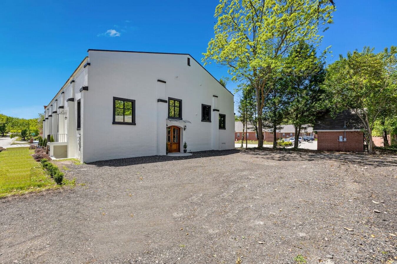 A white building with a front door and windows, surrounded by a gravel driveway and trees.