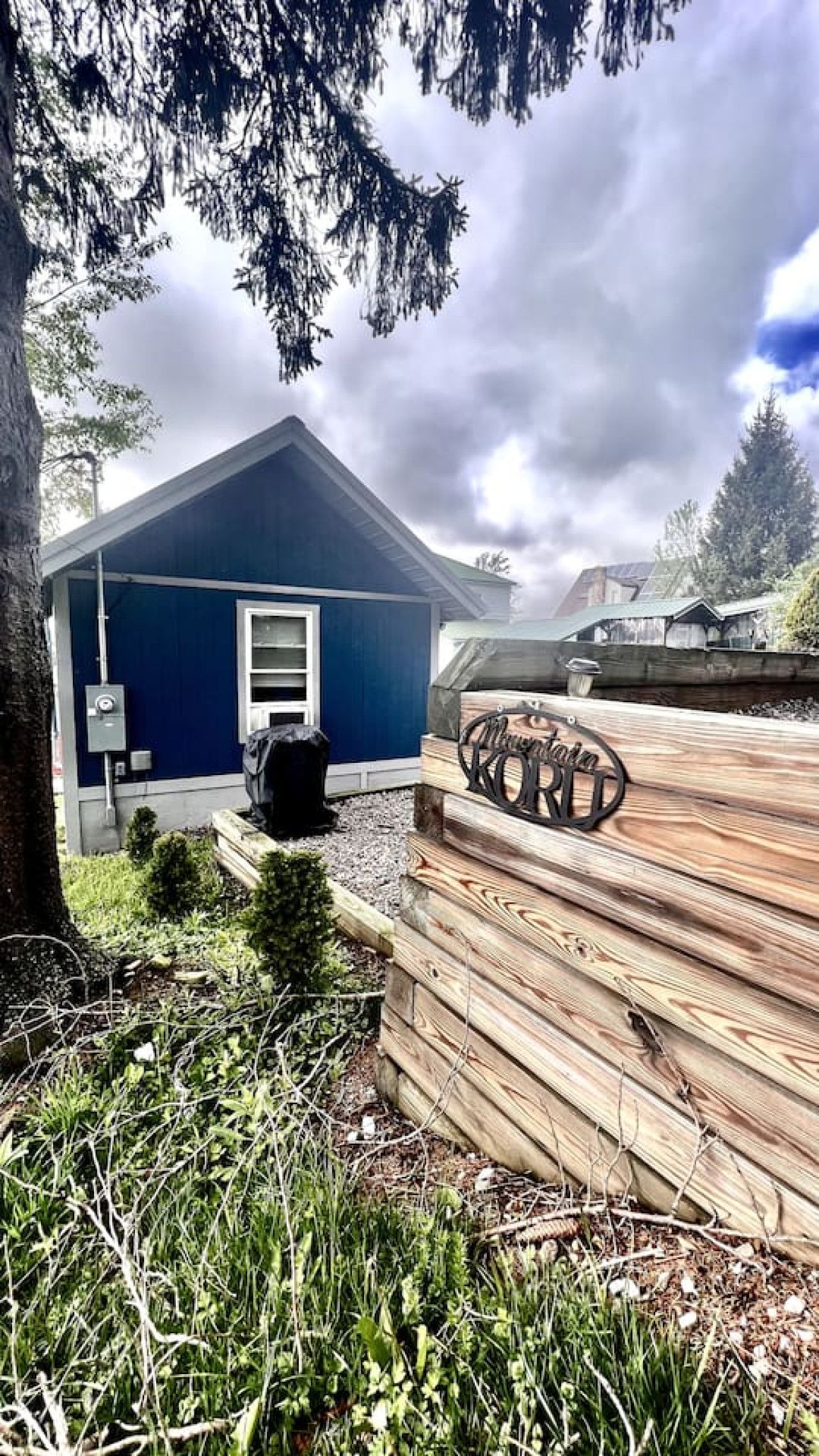 Un petit bâtiment bleu avec une porte et des fenêtres, entouré d’une terrasse en bois et de verdure.