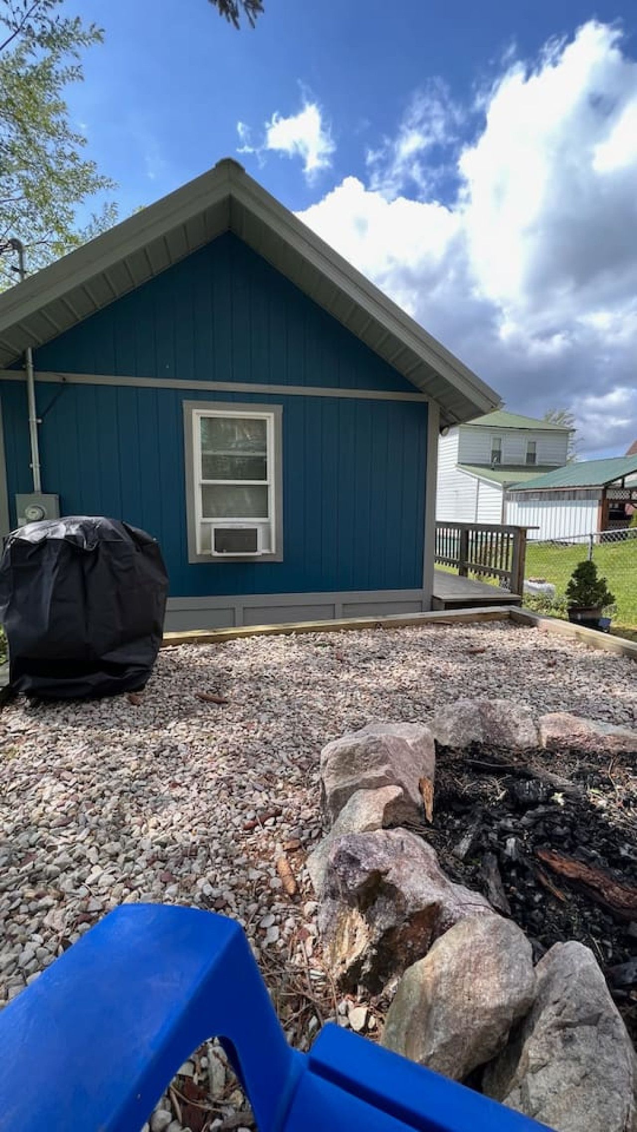 Une maison à l’extérieur bleu, avec une fenêtre et un espace barbecue entouré de pierres, comprenant une chaise bleue.