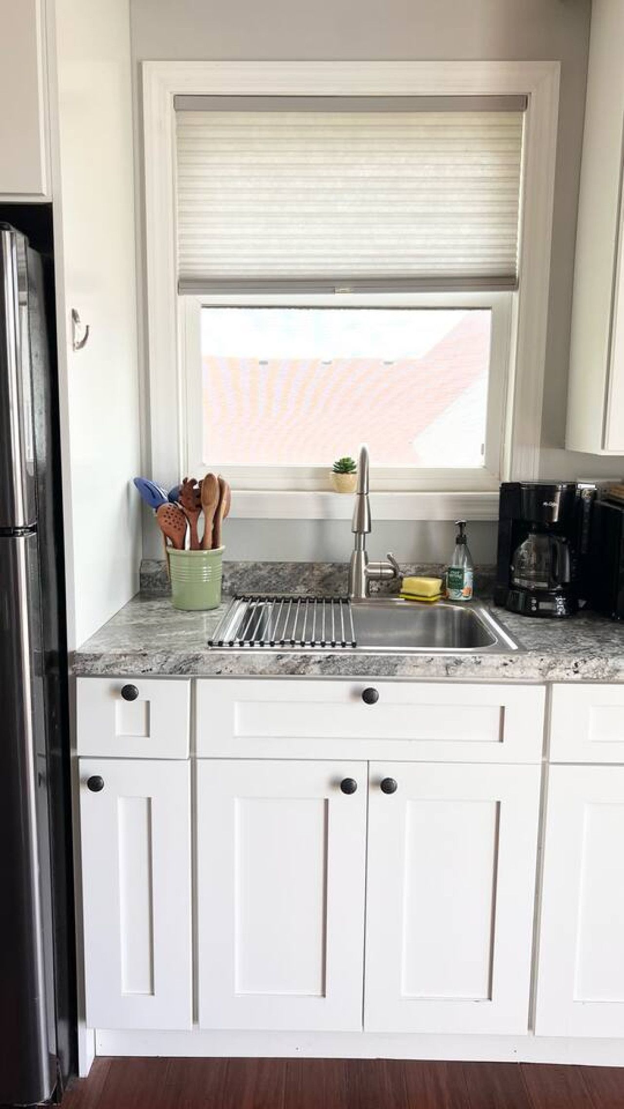 Una cocina con armarios blancos, un lavamanos de acero inoxidable y una ventana con persianas.
