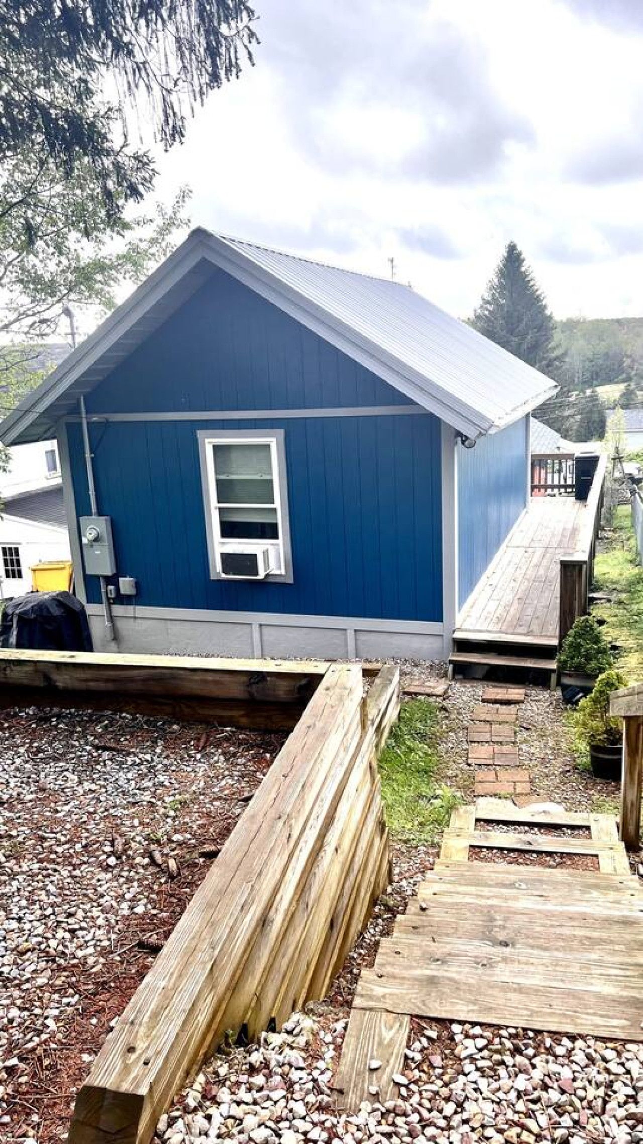 A blue building with a sloped roof, a window, and a door. There is a wooden deck in front of the building.