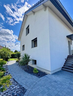 Ein modernes, zweistöckiges Haus mit gepflasterter Auffahrt, einem kleinen Garten mit Steinen und Pflanzen und einem strahlend blauen Himmel mit Wolken.