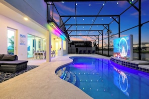 A swimming pool with a curved edge, a covered patio area with seating, and a large digital screen displaying a Ferris wheel.