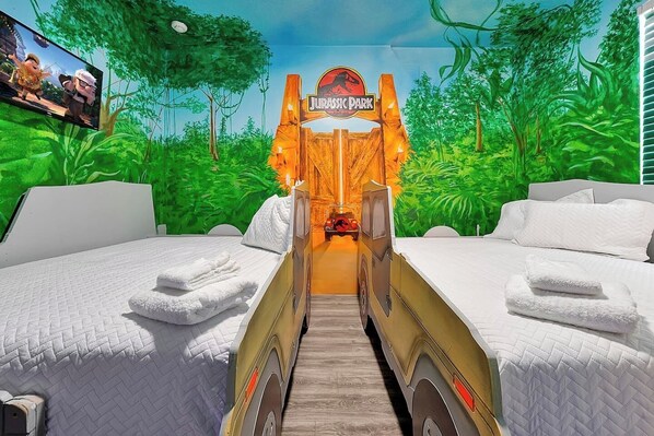 A room with two beds, a jungle-themed mural, a sign reading 'JURASSIC PARK,' and a television screen.